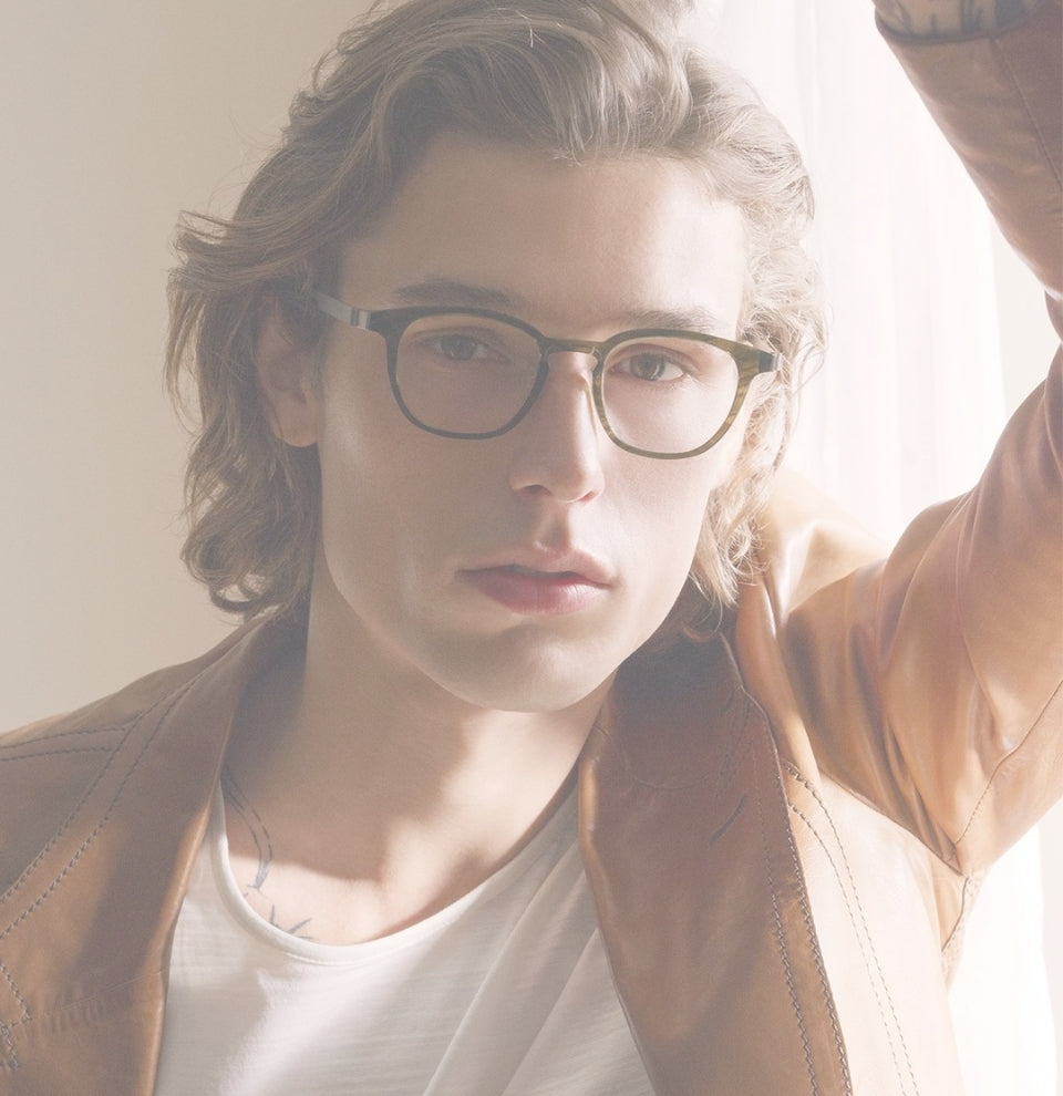 A sun-washed photo of a man in his 20s with collar length, blonde, wavy hair. He wears a white t-shirt and a light brown leather jacket. His left arm is raised and he looks directly at the camera. He is wearing Lindberg horn frames.