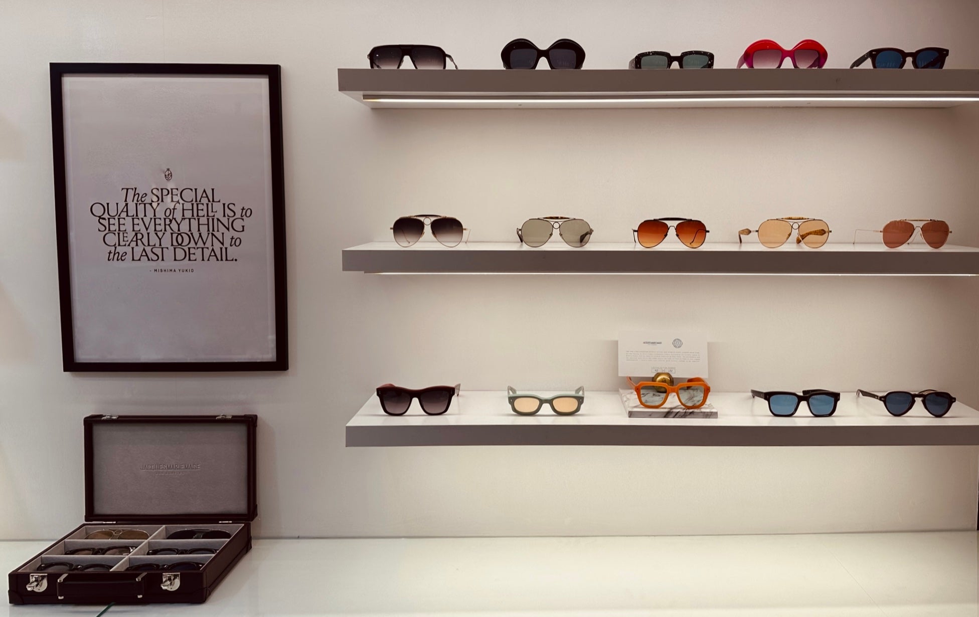 A photo of the back wall of the Dickens Heath practice in warm tones. A selection of frames from Jacques Marie Mage are on display on three parallel shelves. There is a Jacques Marie Mage display box in red leather at the bottom left of the photo. There is a framed photo above it stating "The special quality of hell is to see everything clearly down to the last detail"