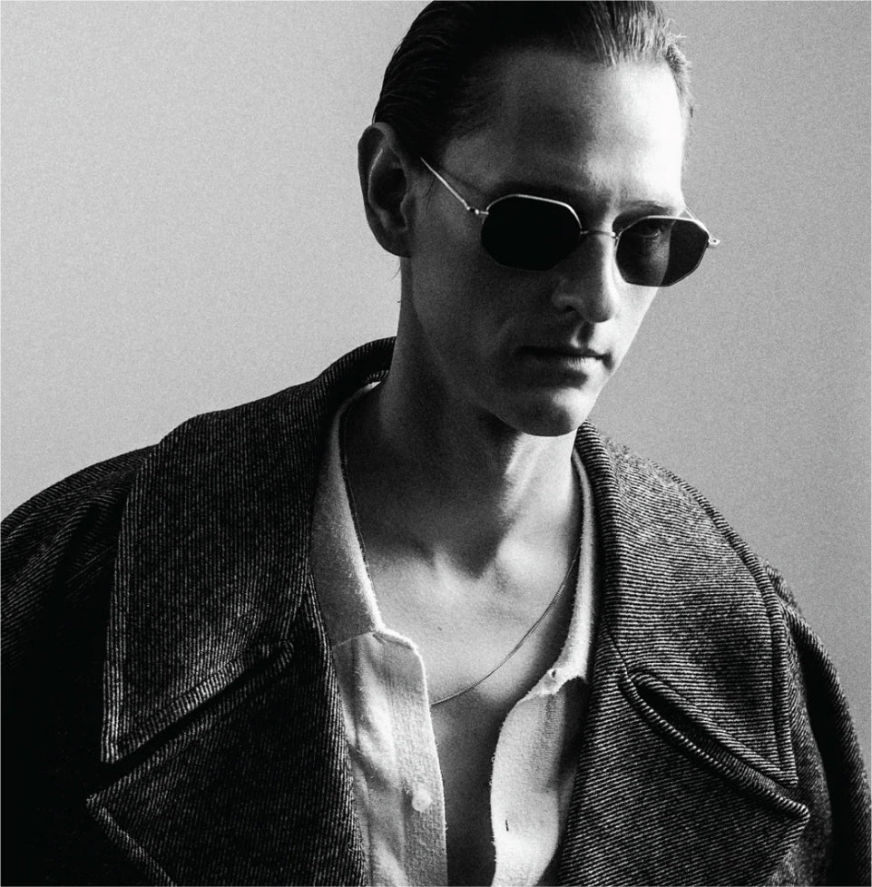 A black and white photo of a man in his 30s with slicked back hair looking down and to his left. He is wearing Ahlem fine-rimmed titanium sunglasses.