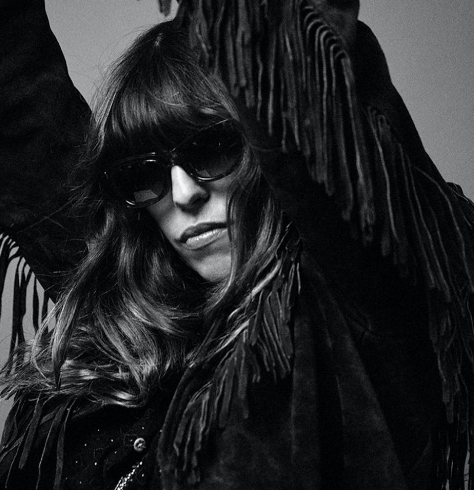 A black and white photo of a woman with long, thick, dark hair and a heavy fringe. Her arms are raised above her head and her shoulders are rotated to the right. Her head is tilted down and to her left but she looks directly at the camera. She is wearing Jacques Marie Mage sunglasses with plastic cat eye frames..