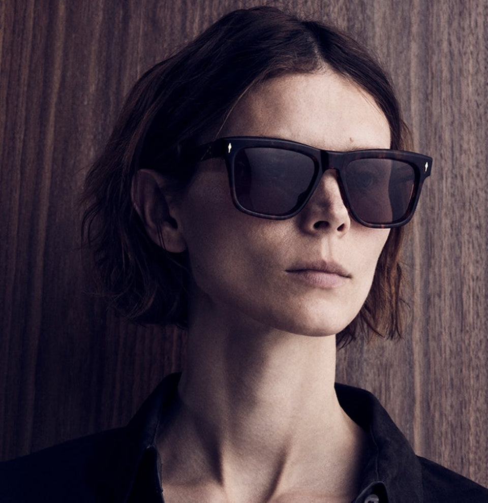 A photo in cool tones of a woman with a short choppy bob, tucked behind her ears, looking to the left. She wears a dark shirt with an open collar. She is wearing Jacques Marie Mage sunglasses with a dark plastic frame.